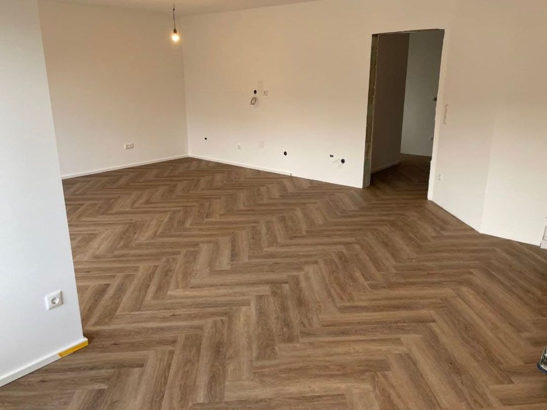 Leerer Wohnraum mit fertig verlegtem Designboden im Fischgrätmuster, helle Wände und eine offene Türdurchsicht